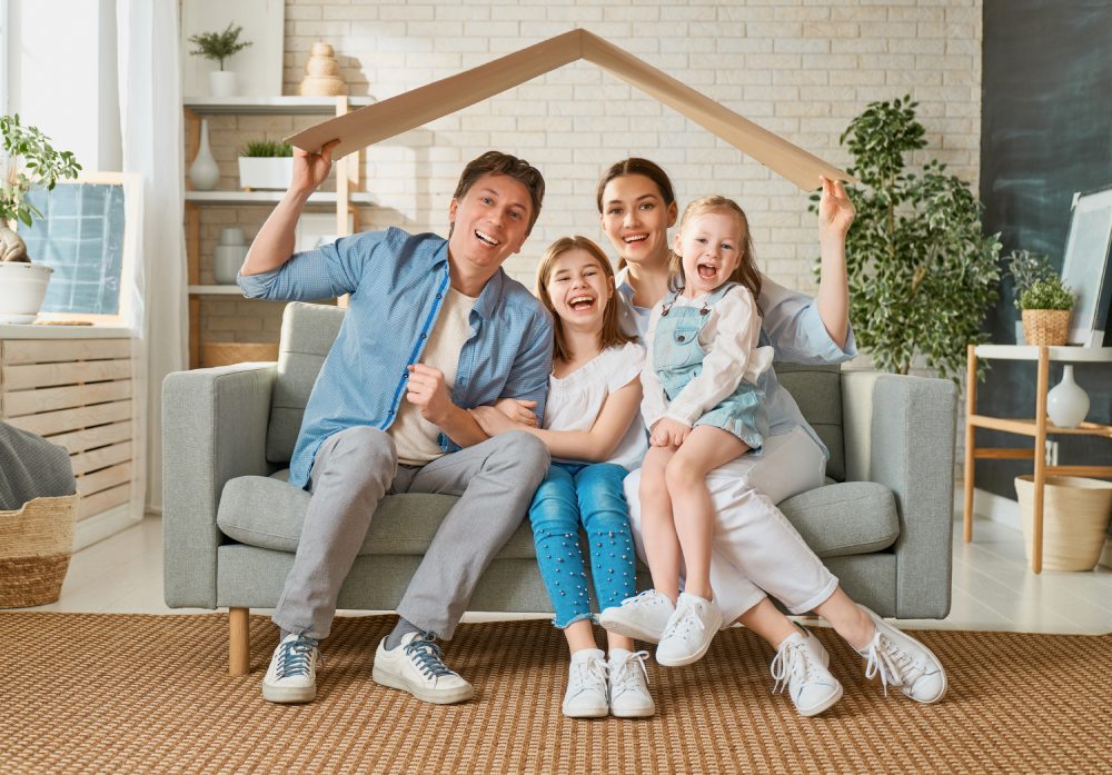 A happy family of four sitting on a couch with a novelty wooden roof being held up inciting they are covered with mortgage insurance